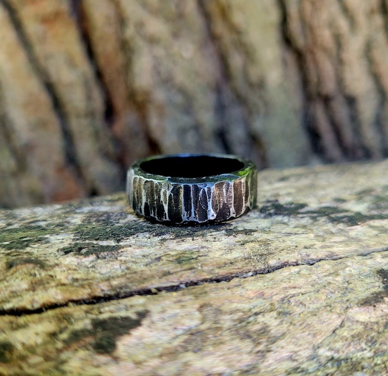 May include: A textured, dark metal ring with a wide band. The ring has a rough, bark-like appearance with vertical grooves and a dark interior. The ring is set against a blurred wooden background.