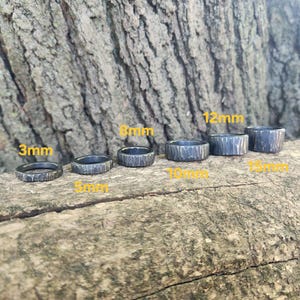 May include: Six dark gray textured metal rings of varying widths, from 3mm to 15mm, are displayed. The rings are arranged on a natural wood surface, with the ring sizes labeled in yellow text.