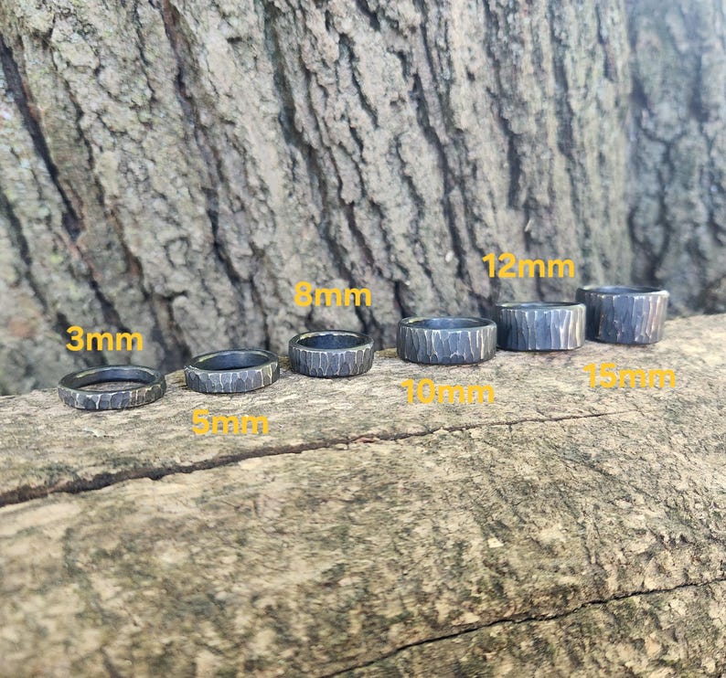 May include: Six dark gray textured metal rings of varying widths, from 3mm to 15mm, are displayed. The rings are arranged on a natural wood surface, with the ring sizes labeled in yellow text.