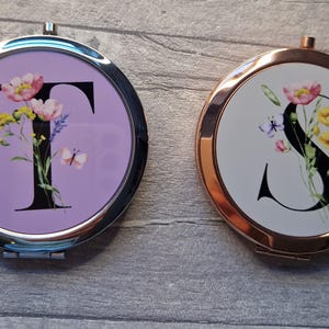 May include: Two compact mirrors with floral designs and letters. The mirror on the left has a purple background with a black letter T and floral accents. The mirror on the right has a white background with a black letter S and floral accents.