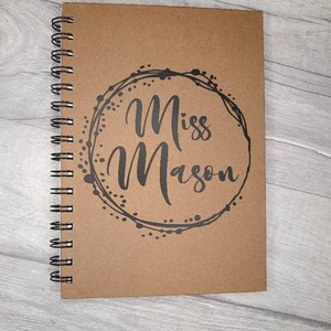 May include: A brown spiral bound notebook with a black circle design and the text "Miss Mason" in a cursive font.