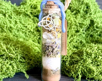 May include: A clear glass bottle filled with layers of herbs, crystals, and sand, sealed with a cork and blue wax. A wooden Celtic knot charm and a small tag are attached. The bottle is surrounded by green moss and dried flowers.