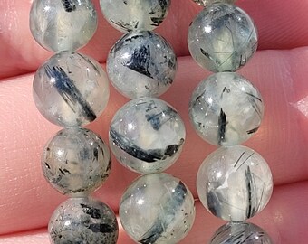 May include: A strand of clear quartz beads with black tourmaline inclusions. The beads are round and have a smooth, polished finish.