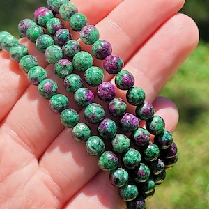May include: Three strands of beaded bracelets. Each bracelet features round beads, mainly green with flecks of deep red and purple. The bracelets are held in a hand, with a blurred green background.