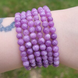 May include: A stack of several bracelets made of round, light purple beads. The beads are strung together, forming a flexible band. The bracelets are worn on a wrist, showcasing the color and texture of the beads.