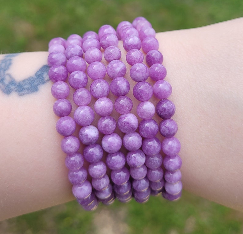 May include: A stack of several bracelets made of round, light purple beads. The beads are strung together, forming a flexible band. The bracelets are worn on a wrist, showcasing the color and texture of the beads.