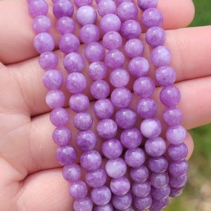 May include: A strand of round, light purple beads. The beads are uniform in size and color, with a smooth, polished surface. The beads are held in a hand against a blurred green background.