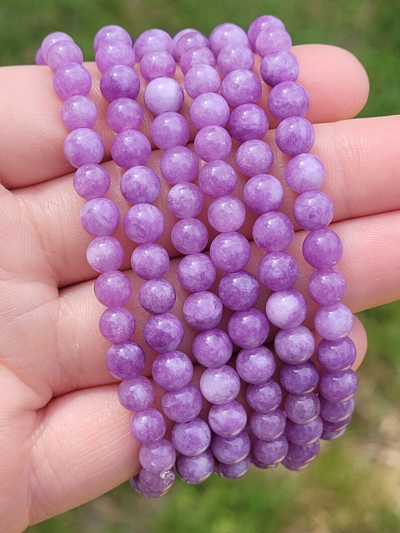 May include: A strand of round, light purple beads. The beads are uniform in size and color, with a smooth, polished surface. The beads are held in a hand against a blurred green background.