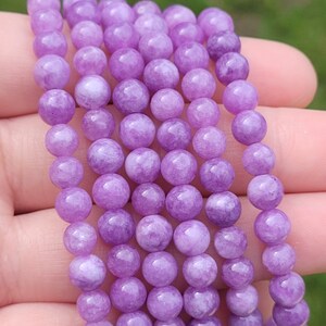 May include: A strand of round, light purple gemstone beads. The beads are smooth and have a slightly translucent appearance. The beads are strung together, creating a necklace or bracelet. The background is blurred, suggesting an outdoor setting.