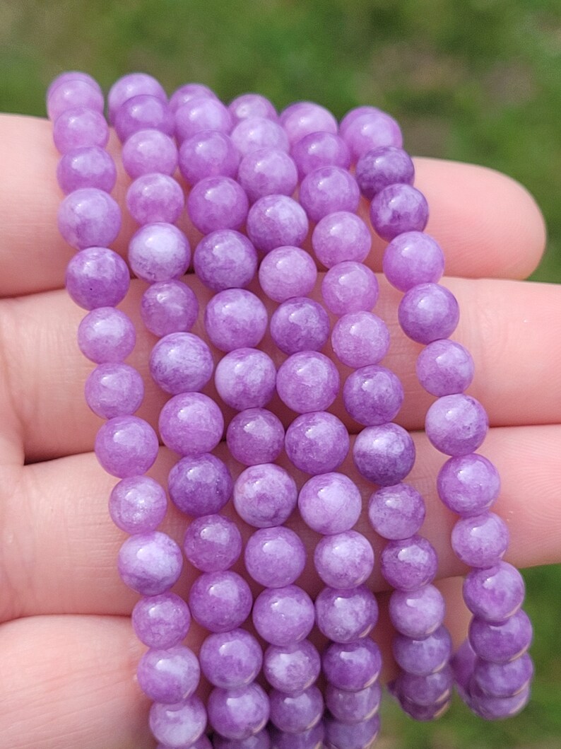 May include: A strand of round, light purple gemstone beads. The beads are smooth and have a slightly translucent appearance. The beads are strung together, creating a necklace or bracelet. The background is blurred, suggesting an outdoor setting.