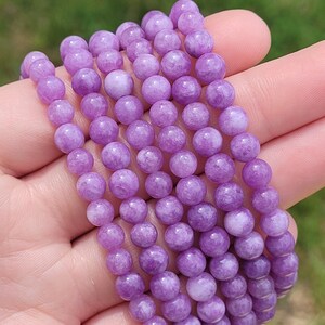 May include: A collection of several beaded bracelets. Each bracelet is made of small, round, light purple beads. The bracelets are held in a hand, with a blurred green background.