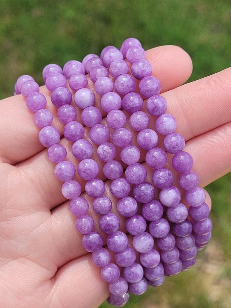 May include: A collection of several beaded bracelets. Each bracelet is made of small, round, light purple beads. The bracelets are held in a hand, with a blurred green background.