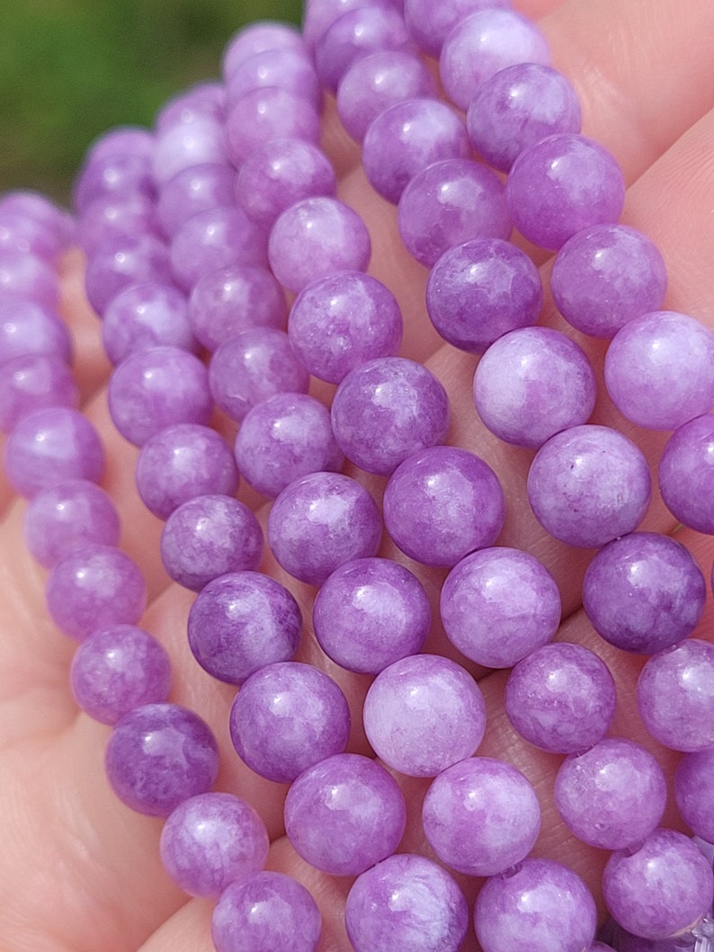 May include: Close-up of several strands of round, light purple beads. The beads have a smooth, polished surface and are strung together. The beads are a soft, muted shade of purple.