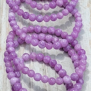 May include: Several strands of lavender-colored beaded bracelets are displayed. The beads are round and have a smooth texture. The bracelets are resting on a light-colored wooden surface.