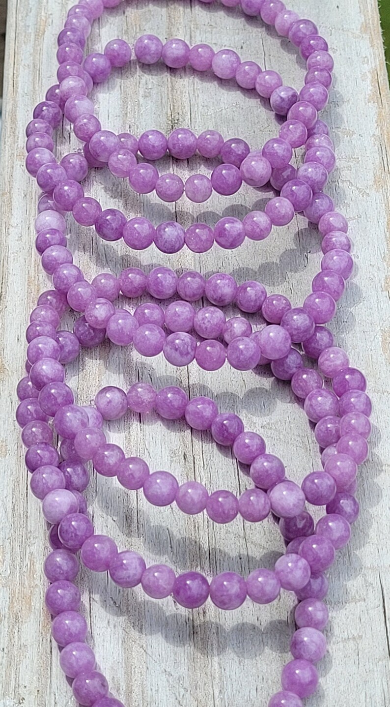 May include: Several strands of lavender-colored beaded bracelets are displayed. The beads are round and have a smooth texture. The bracelets are resting on a light-colored wooden surface.