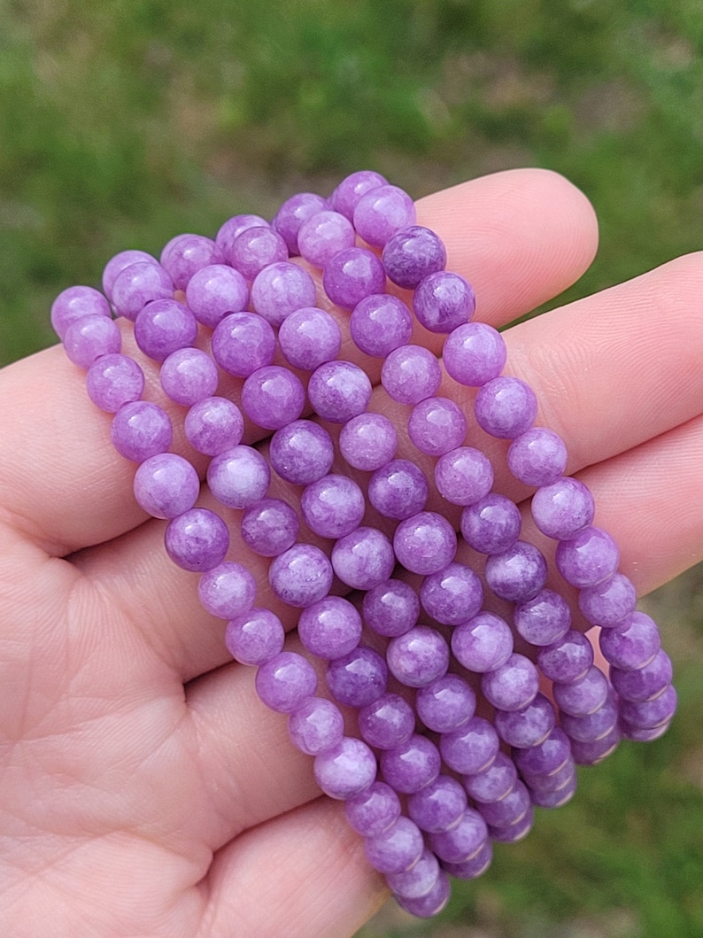 May include: A collection of six purple beaded bracelets. Each bracelet is made of round, polished beads. The bracelets are held in a hand against a blurred green background. The beads have a slight sheen.