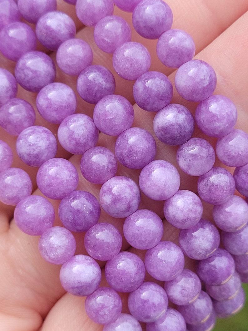 May include: Close-up of a strand of round, light purple beads. The beads have a smooth, polished surface and are strung together. The beads are a soft, pastel purple color.