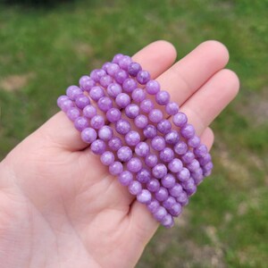 May include: Six strands of round, light purple beads are held in a hand. The beads have a smooth, polished appearance and are strung together. The background is blurred, showing green grass and foliage.