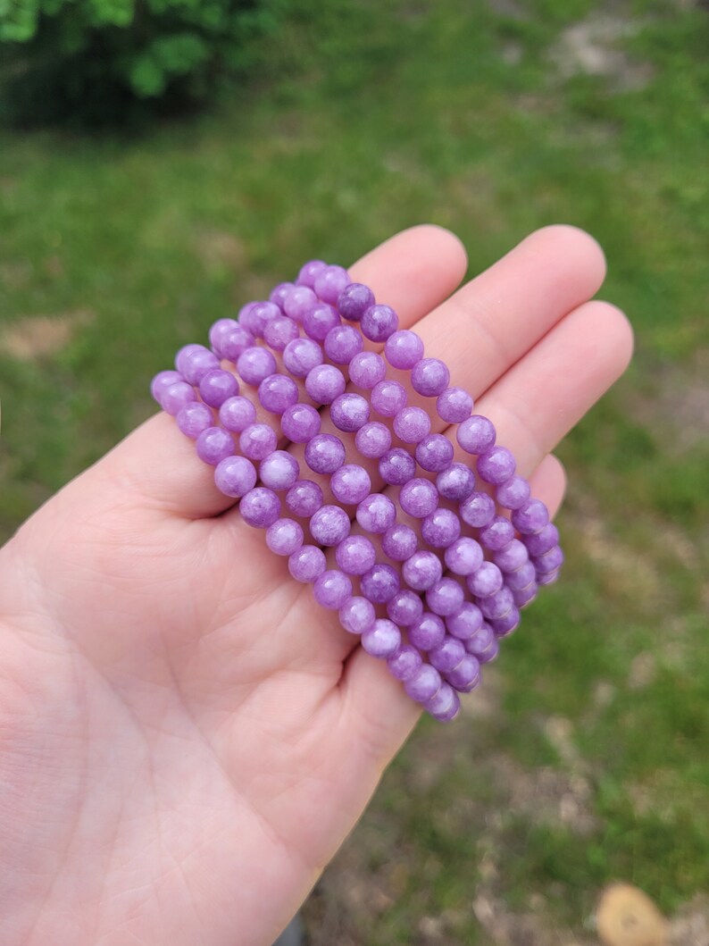 May include: Six strands of round, light purple beads are held in a hand. The beads have a smooth, polished appearance and are strung together. The background is blurred, showing green grass and foliage.