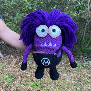 May include: A purple crocheted plush toy with wild purple yarn hair, two large white eyes with black pupils, and a wide mouth showing white teeth. It wears black crocheted overalls with a white M logo and the text MCKNITSCREATIONS on the chest.