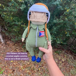 Puede incluir: Muñeco de ganchillo con un mono verde, zapatos azules y un sombrero blanco y naranja. El muñeco tiene una cara bronceada y trenzas naranjas. Un pequeño parche de la bandera puertorriqueña está en el mono. El texto de la imagen dice: "PDF Pattern, on how to crochet sapito concho, this is not a finish product."
