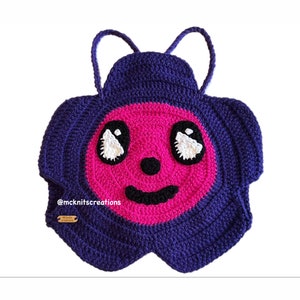 Op de afbeelding: Een handgemaakte, gehaakte tas in een grillig ontwerp. De tas heeft een paarse buitenlaag met een roze gezicht en zwart-witte ogen. De tas heeft twee paarse bandjes en een lachende mond. De tekst "@mknitscreations" is zichtbaar.