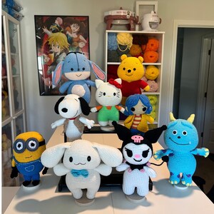 May include: A collection of handmade crochet dolls featuring various cartoon characters, including Winnie the Pooh, Hello Kitty, Snoopy, and a Minion. The dolls are in various colors and sizes, displayed on a white surface.