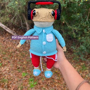 May include: A handmade crochet frog doll wearing a light blue jacket, red trousers, and blue shoes with white star accents. The frog has a tan head, black headphones with red accents, and a white face mask. The text "PDF English Pattern" is visible.