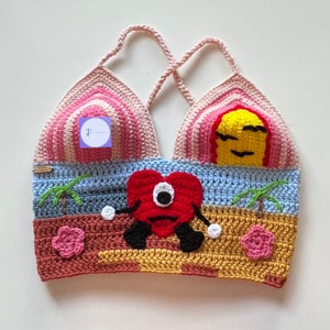 Op de afbeelding: Een gecrochete crop top met een strandontwerp. De top heeft een roze, gele en blauwe zonsondergang, palmbomen en een rood hartvormig monster met één oog.