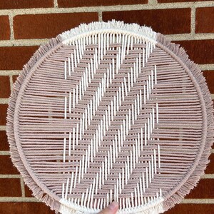 May include: A round macrame wall hanging with a pink and white geometric pattern. The hanging has a fringe border.