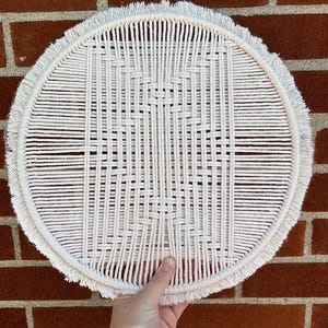May include: A white macrame wall hanging with a fringe border. The hanging is circular and has a woven pattern.