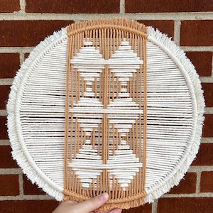 Könnte beinhalten: Ein runder, gewebter Wandbehang mit geometrischem Design. Das Stück hat einen cremefarbenen Außenring mit Fransen, eine tanfarbene Mitte und ein weißes geometrisches Muster. Der Wandbehang wird vor einer roten Backsteinmauer gehalten.