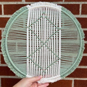 Puede incluir: Un tapiz de macramé redondo con un diseño blanco y verde salvia. El tapiz tiene un borde de flecos.