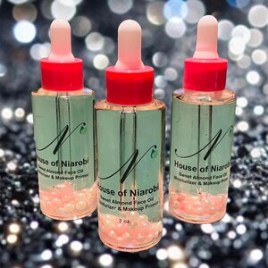 Puede incluir: Tres botellas de vidrio de aceite facial de almendras dulces y prebase de maquillaje. Las botellas son transparentes y tienen un gotero rosa. Las botellas están etiquetadas con "House of Niarobi" y tienen un logotipo de hoja verde. Las botellas están llenas de un líquido transparente y tienen pequeñas cuentas rosadas en el fondo.