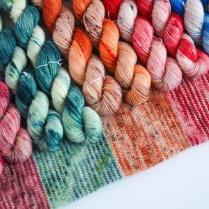 Könnte beinhalten: Nahaufnahme einer Vielzahl von bunten Garnknäueln, die in einem Regenbogenmuster angeordnet sind. Das Garn wird auf einem gestrickten Stoff mit roten, grünen, blauen und orangen Streifen gezeigt.