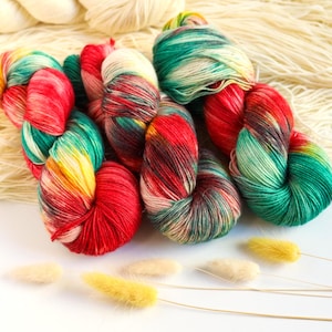 Può includere: Tre matasse di filato tinto a mano caratterizzate da una tavolozza di colori variegati di rosso vibrante, verde smeraldo e giallo dorato, appoggiate su uno sfondo di matasse di filato color crema non tinto e steli d'erba decorativa.