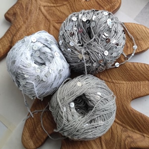 May include: Three balls of yarn in shades of gray and white, adorned with silver sequins. The yarn is arranged on a wooden, leaf-shaped tray. The yarn is suitable for knitting or crochet projects.