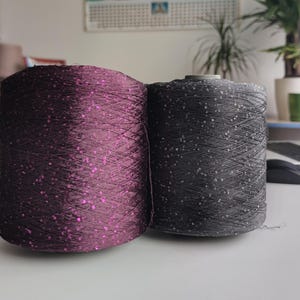 May include: Two spools of yarn. One is a deep purple color with pink sequins, and the other is dark gray with silver sequins. The yarn is tightly wound, and the spools are side by side. The yarn is likely for knitting or crochet projects.