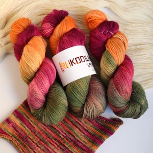 Può includere: Tre matasse di filato variegato con una sfumatura di arancione, magenta e verde oliva. Un'etichetta di carta bianca con il testo RUKODILNYA è avvolta attorno alla matassa centrale. Sotto si trova un campione lavorato a maglia a righe orizzontali.