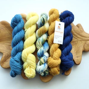 May include: Five skeins of yarn in blue, yellow, variegated blue and white, mustard, and dark blue, arranged side by side. A white rectangular tag with printed text is attached to the mustard yarn. The yarn rests on a wooden leaf-shaped tray.