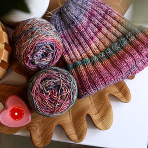May include: A handmade knit hat in shades of purple, pink, and orange sits next to two balls of yarn with similar colors. The yarn and hat are on a wooden tray with a heart-shaped candle holder.