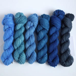 Peut inclure: Six écheveaux de fil bleu dans différentes nuances, du clair au foncé. Le fil est torsadé serré, révélant la texture des fibres. La gamme de couleurs comprend le bleu ciel, le sarcelle et le marine, créant un effet de dégradé.