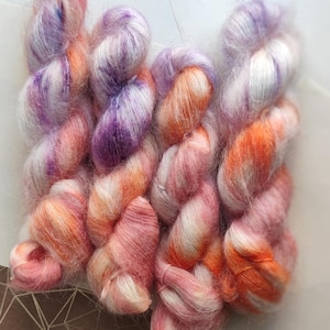 Puede incluir: Primer plano de madejas de hilo coloridas en tonos rosa, naranja, morado y blanco. El hilo parece suave y esponjoso, con un ligero brillo. Las madejas están dispuestas en forma rectangular, lo que sugiere un proyecto de manualidades o material para tejer o hacer ganchillo.