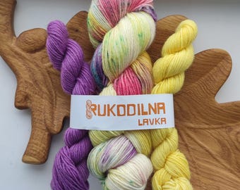 May include: Three skeins of yarn bundled together with a white paper label reading RUKODILNA LAVKA. The left skein is solid purple, the center skein is variegated with cream, pink, yellow, and green speckles, and the right skein is solid yellow.