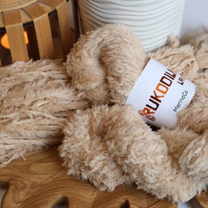 May include: Close-up of beige yarn with a fluffy texture, labeled "Rukodilka MerinoCo." The yarn is arranged with other strands on a wooden surface. A decorative wooden lantern and a white patterned container are in the background.