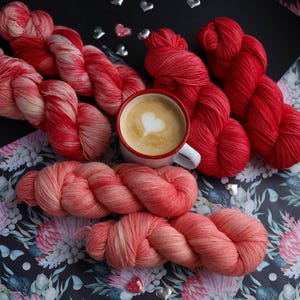 Può includere: Una vista dall'alto mostra gomitoli di filato rosso e rosa, una tazza di caffè con un motivo a forma di cuore nella schiuma e decorazioni a forma di cuore. Il filato è disposto su una superficie a motivi floreali, creando una scena calda.