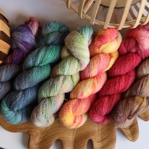 May include: A selection of vibrant yarn skeins in hues of purple, teal, green, yellow, red, and brown. The yarn is displayed on a wooden decorative piece. A woven basket and a white surface are in the background.