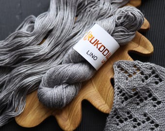 May include: A skein of gray yarn with a white label that reads "Rukodil Lino" sits on a wooden cutting board. Loose strands of yarn are arranged to the left. A knitted swatch with a diamond pattern is on the right. The background is dark.