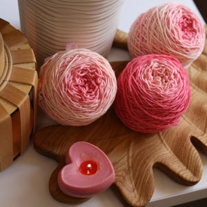 May include: A wooden leaf-shaped tray holds three balls of pink and red yarn. A heart-shaped candle with a lit flame sits on the tray. A woven lantern and a decorative vase are also present, creating a cozy, craft-themed scene.