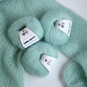 May include: Three balls of light teal Lana Gatto Silk Mohair Lux yarn with white labels. The yarn is fluffy and soft, with a slight sheen. The background includes a knitted fabric in the same color.
